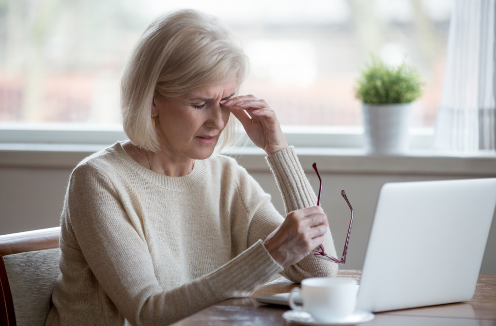 Senior woman rubbing irritated dry eyes with one hand in front of laptop