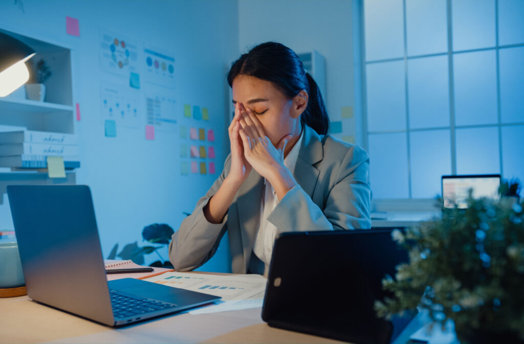 A professional-looking woman rubbing her eyes while working in a dimly lit room.