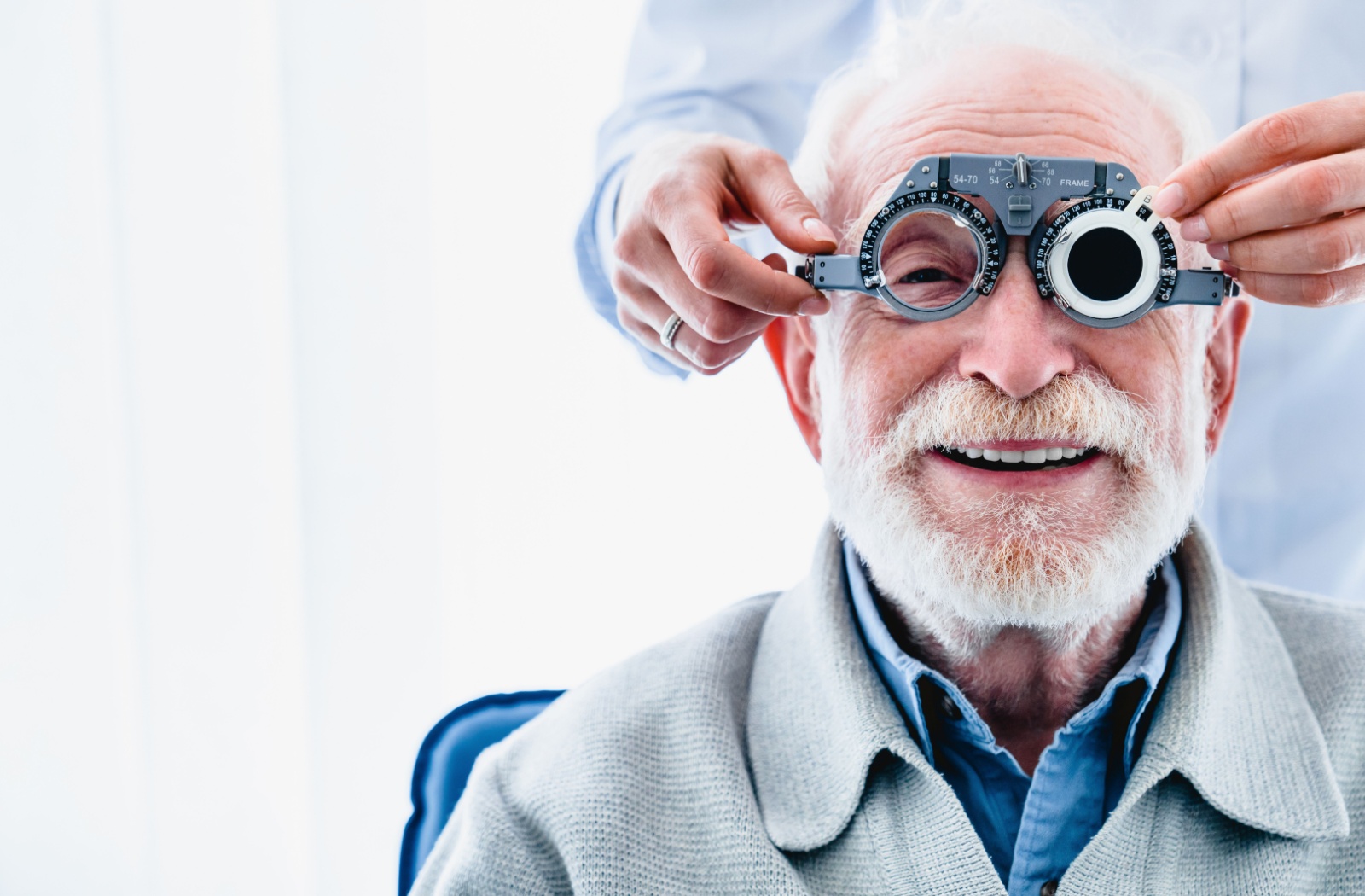 An older adult smiling during an eye exam while an out-of-frame optometrist uses phoropter glasses to diagnose his refractive error.
