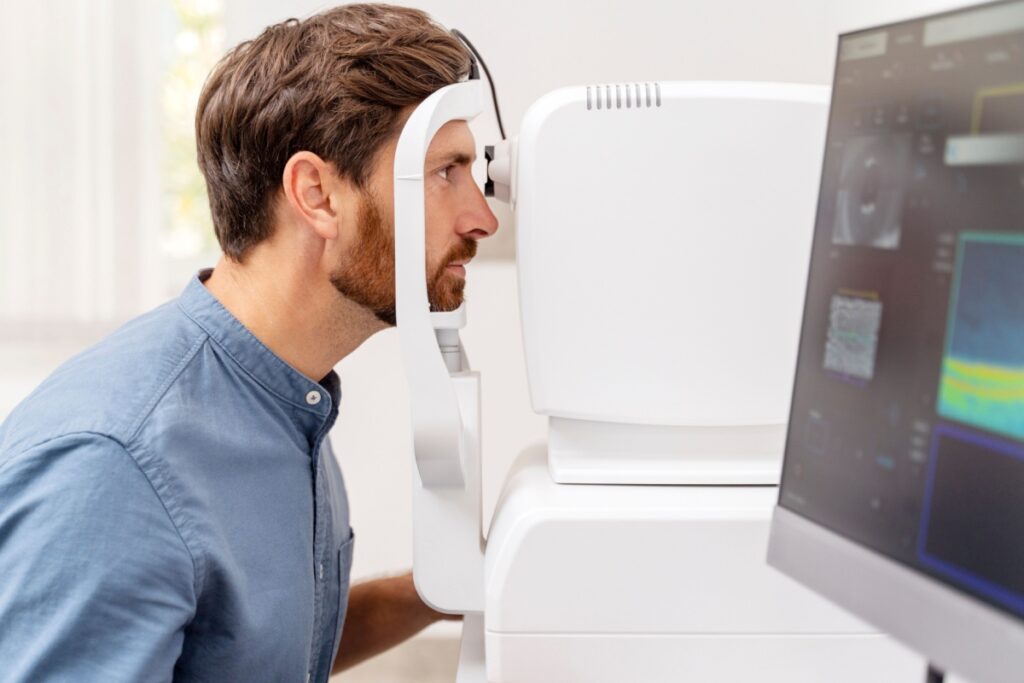 Man in denim shirt positioning his face against a white optical diagnostic device while looking at a computer monitor displaying eye exam results during a comprehensive eye examination.