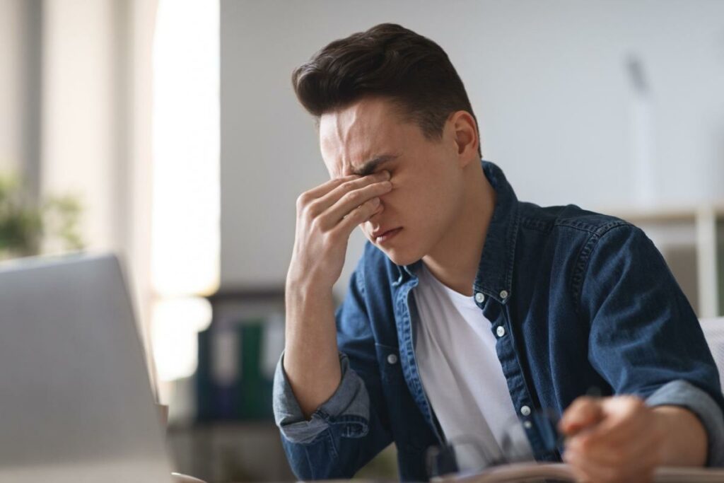 Young man rubbing his eyes while working at a laptop computer.